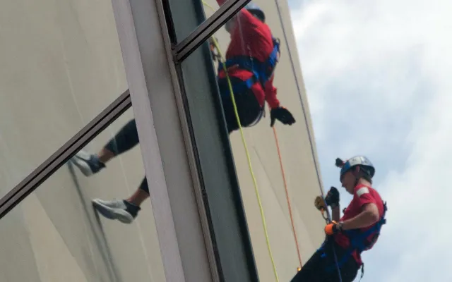 Window cleaner in red and black gear, reflected in glass building facade.
