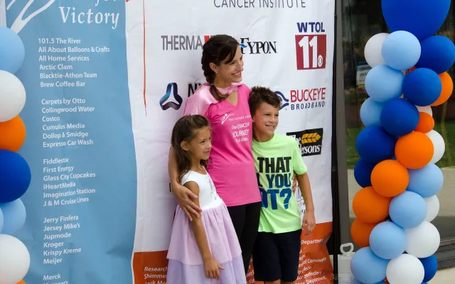 Woman and two children pose at an event, surrounded by colorful balloons.