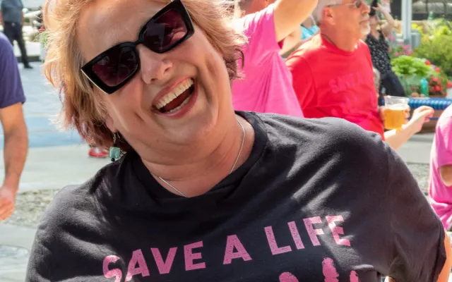 Woman wearing sunglasses and a humorous T-shirt, smiling outdoors with others nearby.