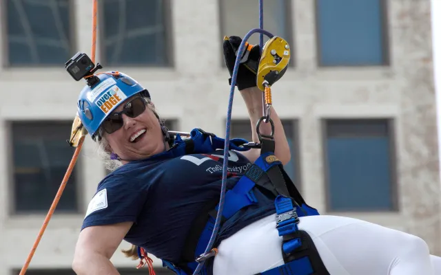 A person wearing climbing gear and helmet, happily rappelling from a building.