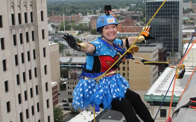 Two climbers in costumes rappel down a tall building with a city view behind them.