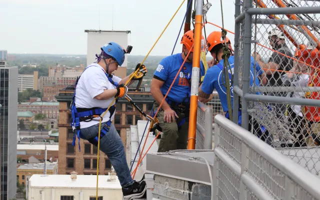 Person in harness rappelling down a building, assisted by workers in helmets. Urban background.
