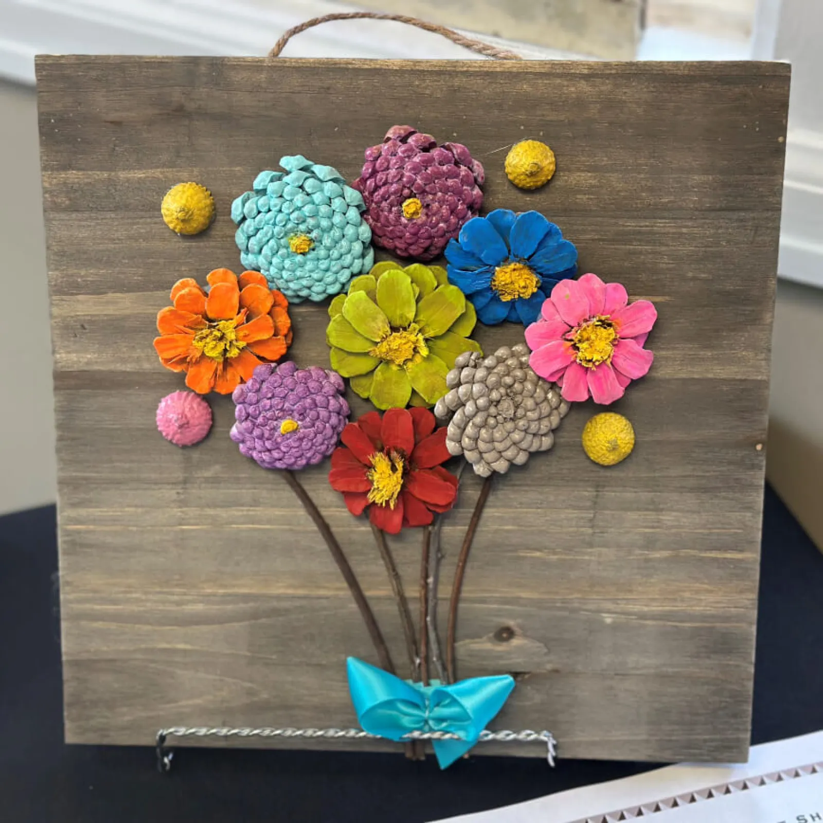 Colorful flower artwork on wooden canvas with a small blue paper boat.