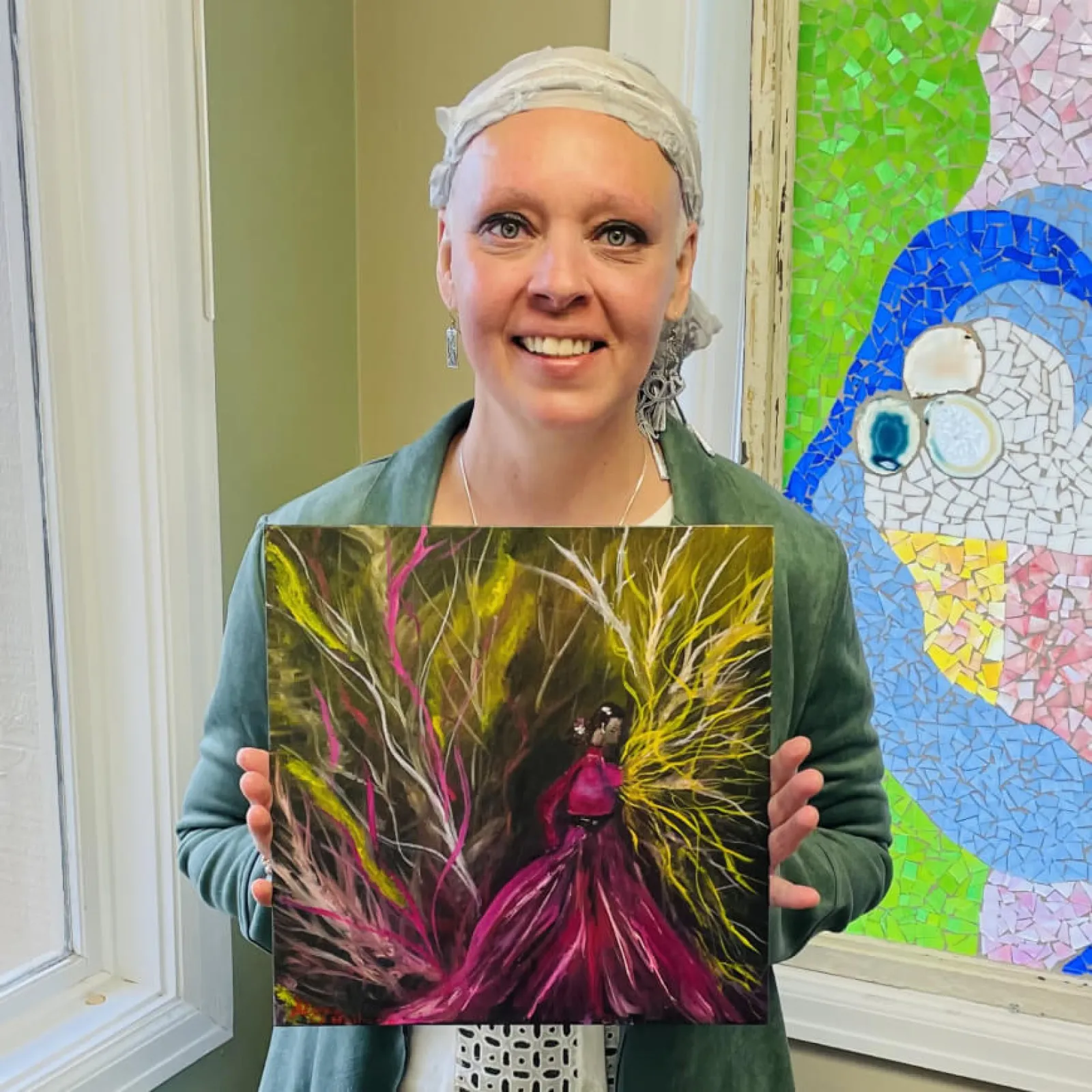 Woman in headscarf holding vibrant painting, colorful mosaic behind her.