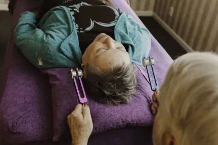 Person lying on a purple mat with tuning forks held near their ears.